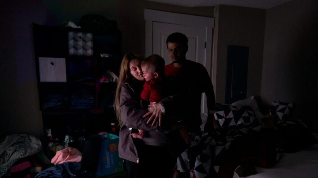 A woman holds a small child as a man stands behind her. The photograph is dark, as if there wasn't much light in the room.