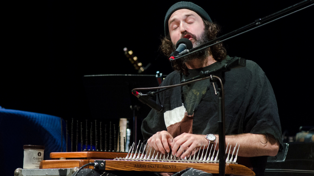 A man with facial hair wearing a beanie sings into a mic while playing the mbira.