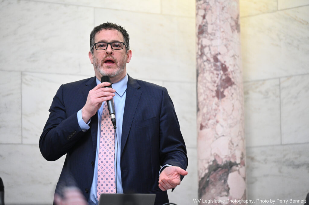 Man in suit, glasses and beard speaking into microphone in large room