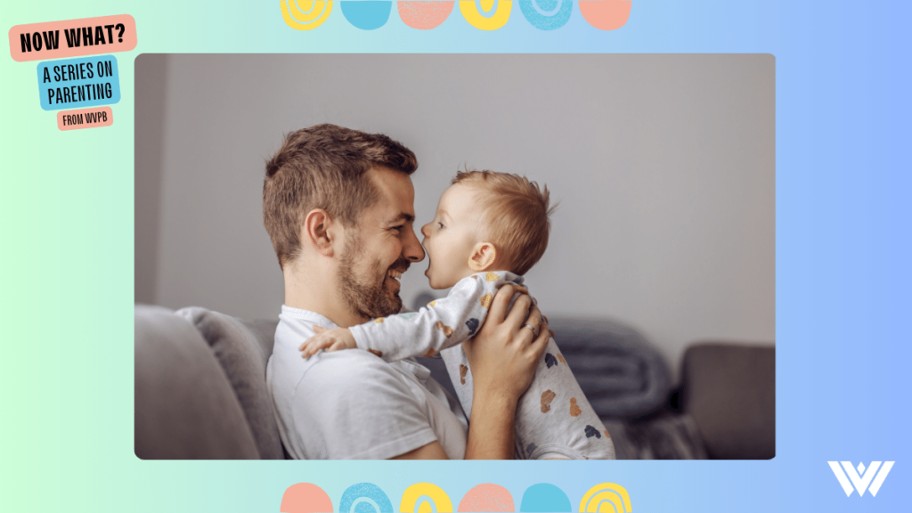 A man in a white shirt with a light beard sits on a grey couch while holding an infant facing him. The infant, wearing a white onesie with a multicolored pattern of animal outlines is attempting to put the man's nose into his mouth. The image is bordered by a blue gradient with pastel colored circles above and below. In the top left of the frame are the words "Now What? A Series On Parenting" and in the bottom right is the WVPB logo.
