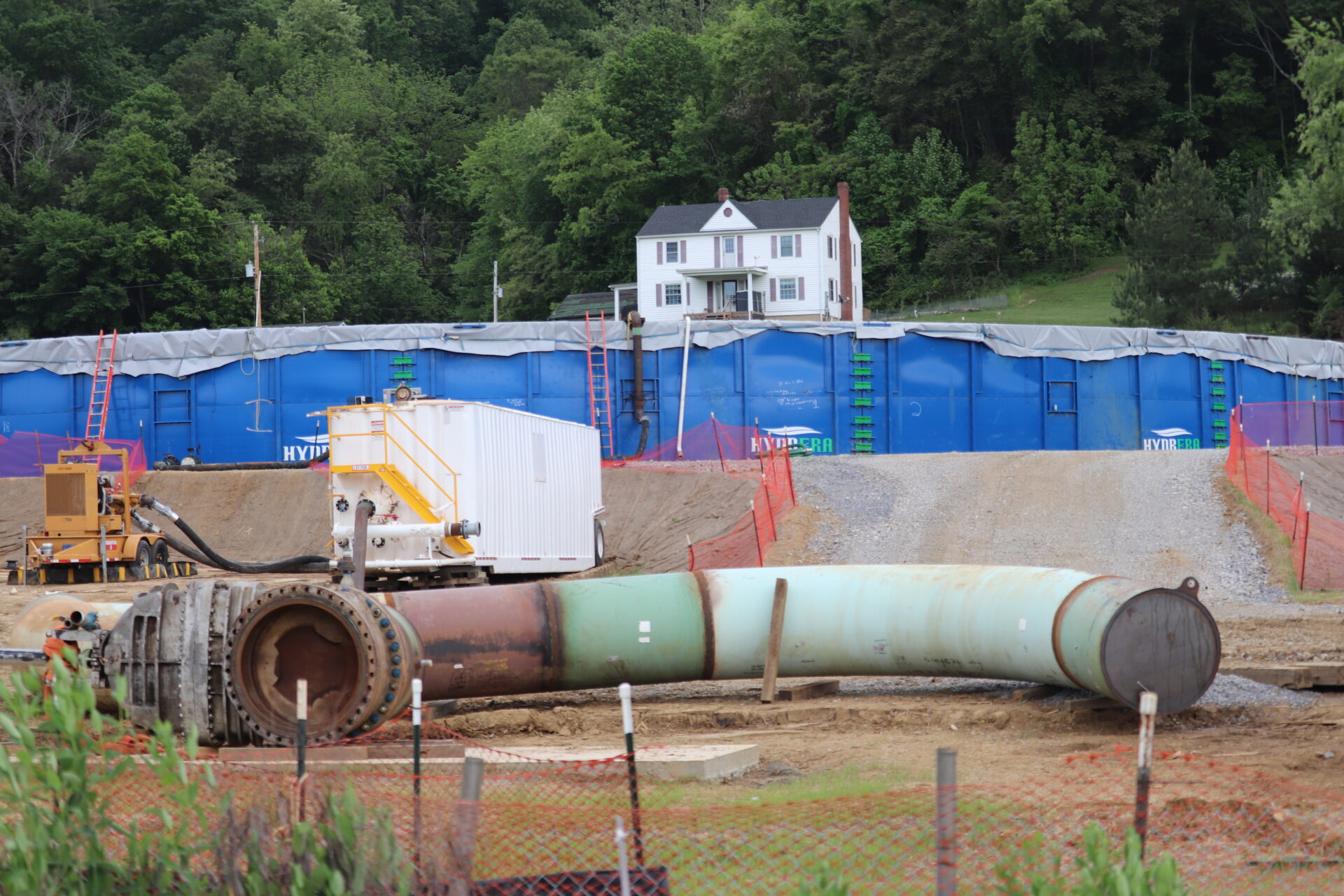 A large section of green pipe lies on the ground in front of a large blue water storage tank and a residence.