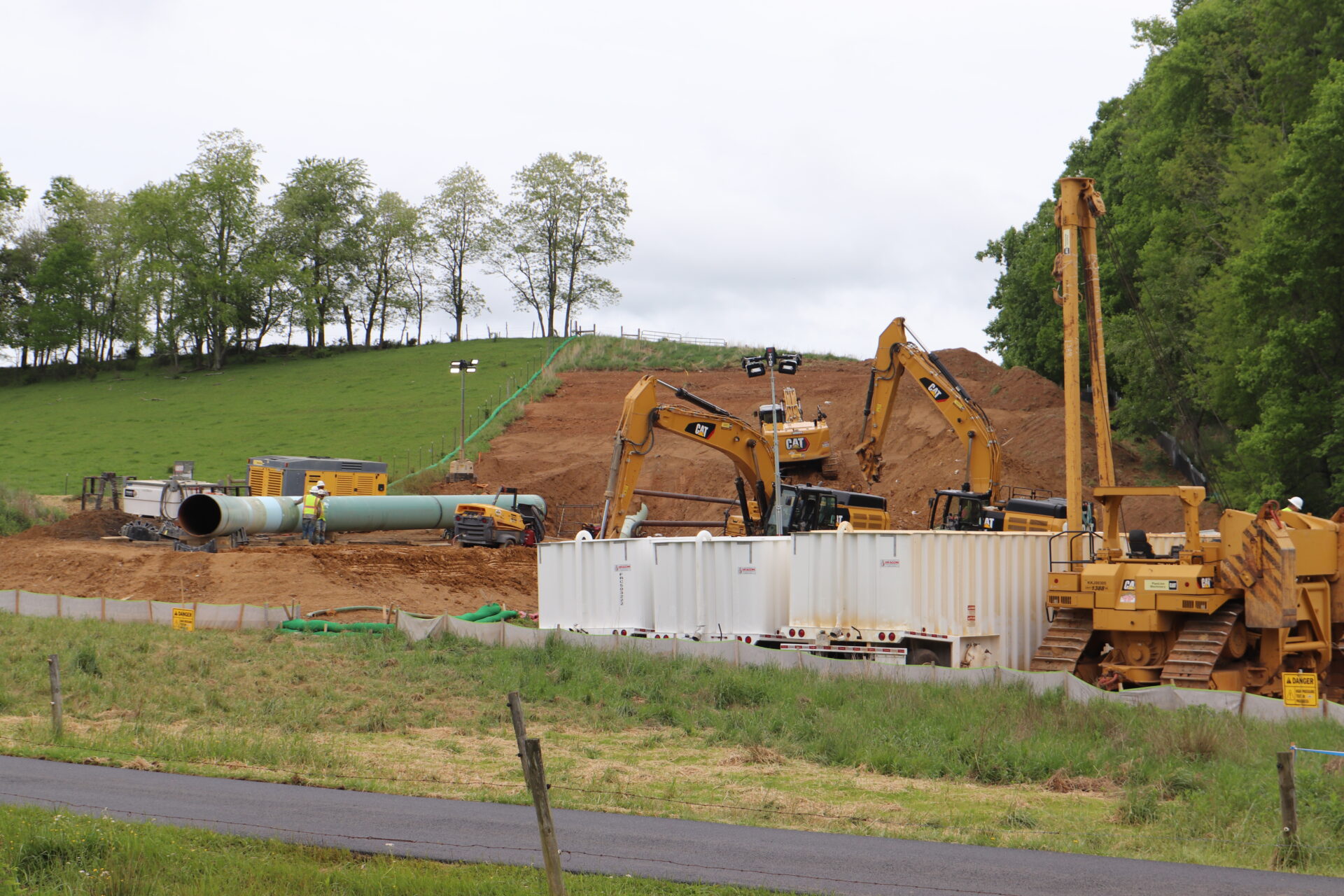 Exposed dirt on a hillside with sections of pipe, storage containers and construction machinery surrounding it under an overcast sky.