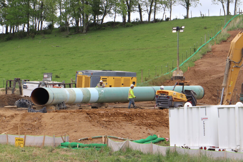 A section of green pipe lays in exposed red dirt next to a green slope with workers next to it.