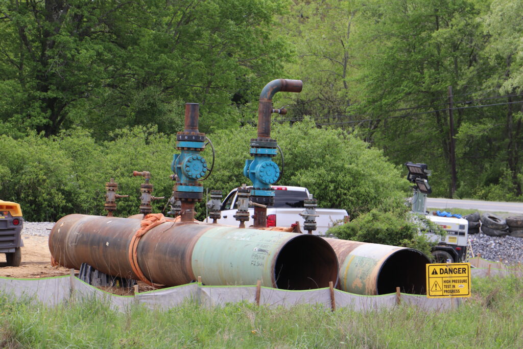 Two sections of pipe rest on the ground as a yellow sign warns people of pressure testing for a natural gas pipeline.