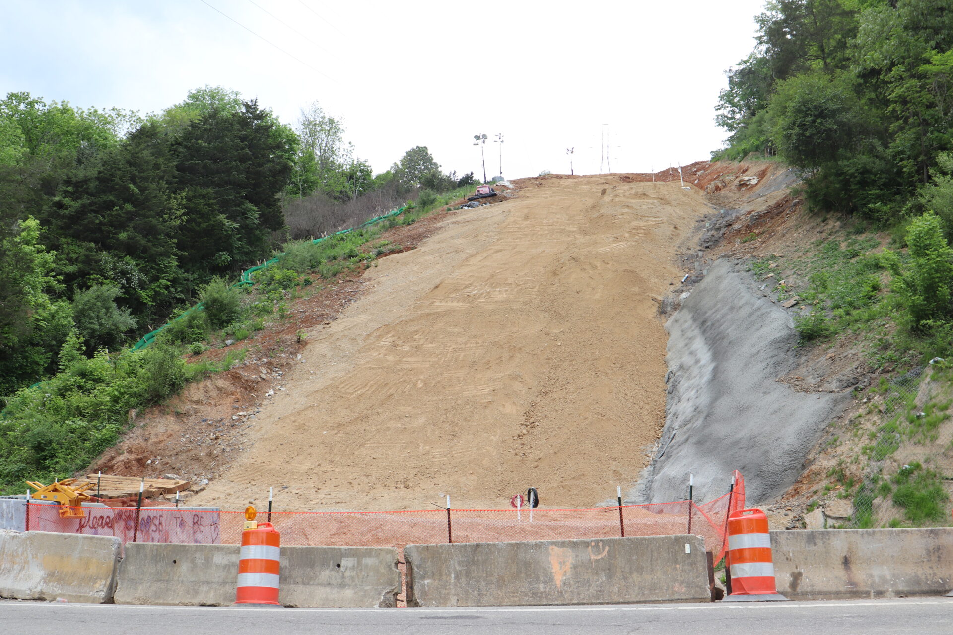 A steep earthen slope is viewed from a county road with concrete barriers and two orange and white construction barrels at the bottom.