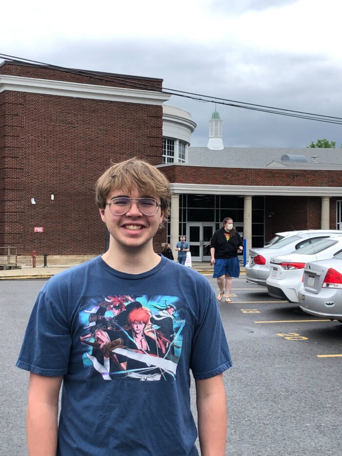 A young man with glasses smiles for the camera. He wears an anime ts-shirt and stands in front of a school.