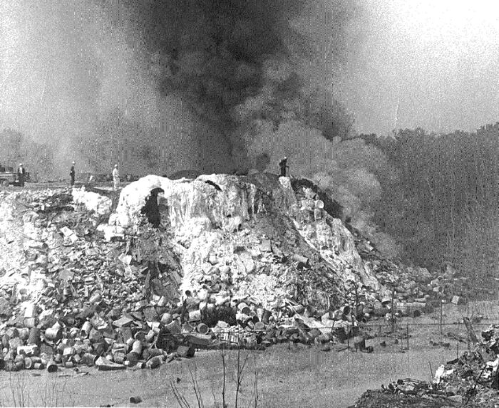 A pile of industrial waste burns and emits a cloud of dark smoke in a black and white photo.