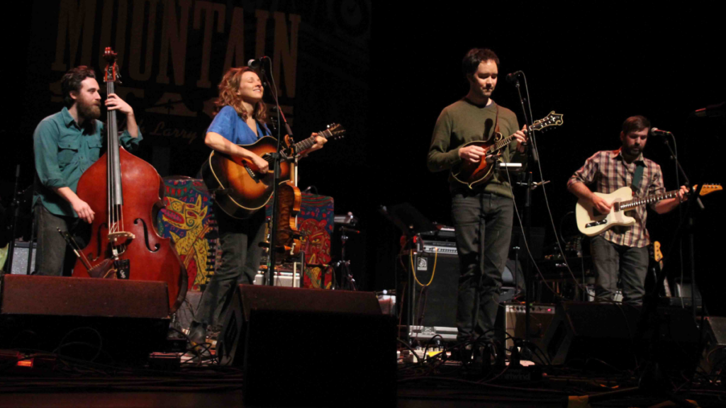 Four musicians are seen performing on a stage. One plays a cello, one plays a mandolin, and the other two play guitars.