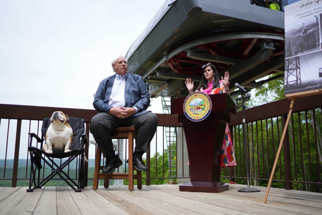 Lady in flowery dress speaking at podium with large man and fat dog next to her