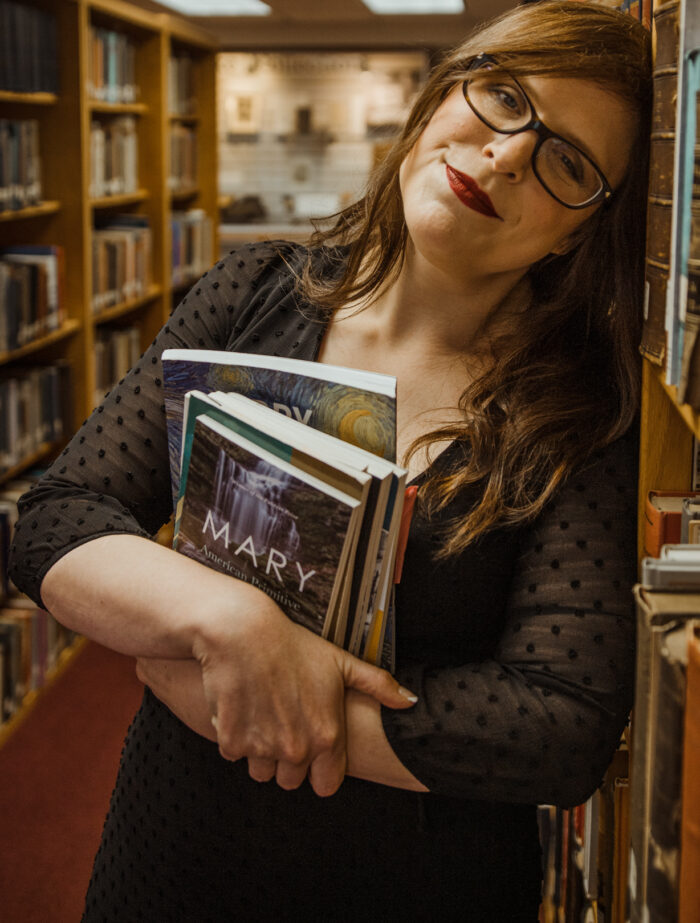 A photograph of a woman with brown hair and glasses leaning against a bookshelf. In her arms, she holds a small pile of books.