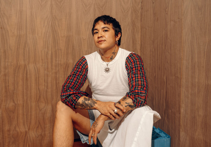 A young man poses for a photo. He has dark hair, wears a long-sleeved shirt that is white with plaid arms. He has a tattoos on his arms.