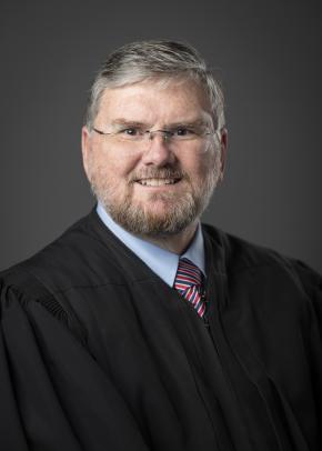 A man in glasses and a judge's cloak sits and looks directly into the camera for a headshot photo.