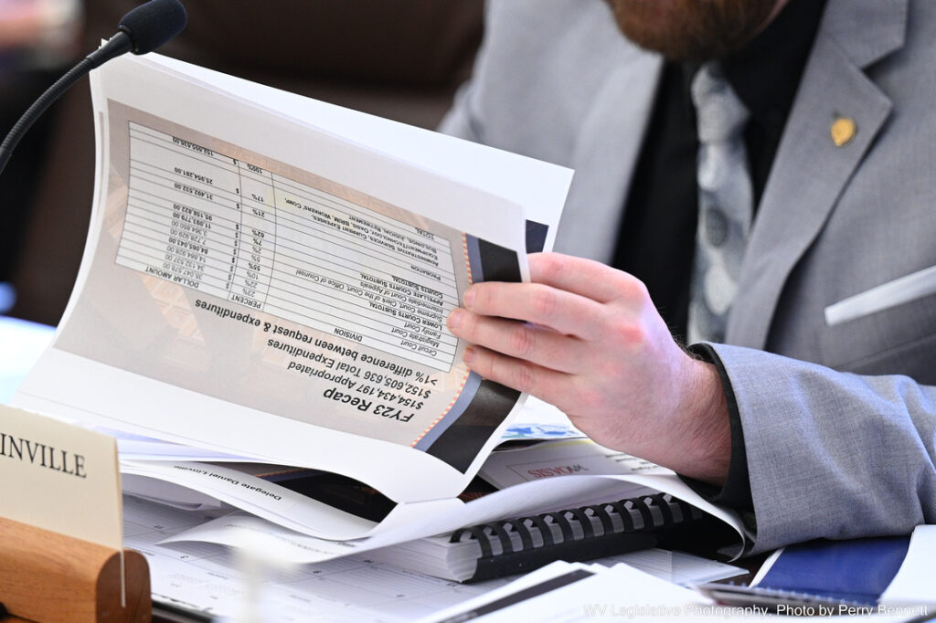 A man in a suit and tie rifles through a packet of papers. The top page he is flipping through has a spreadsheet labeled "Fiscal Year 2023" and several rows of financial information.