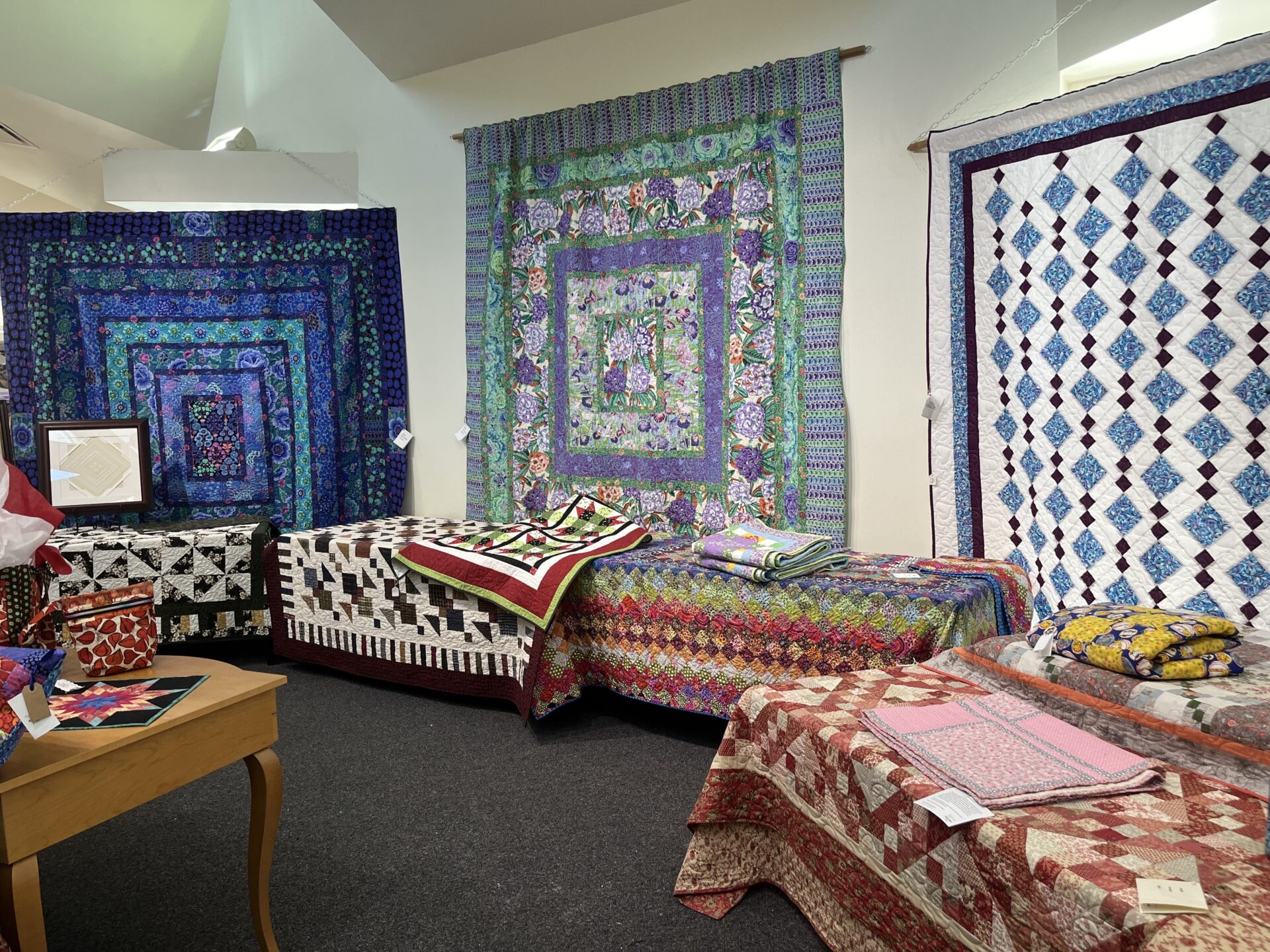 Colorful quilts are spread across several tables placed along the walls of a carpeted room. Behind the tables, additional quilts hang from the walls.