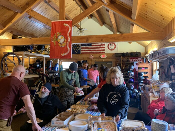Women behind tables ladle food onto the plates of a line of men in a log wood room. The men are lined up along the tables with plates in hand, some of them talking and others focusing on the food before them. The room has an angled roof with exposed wood beams. An American flag, Marine Corps flag and West Virginia University flag are on display. Around the room, snowboards and ski equipment are stored.