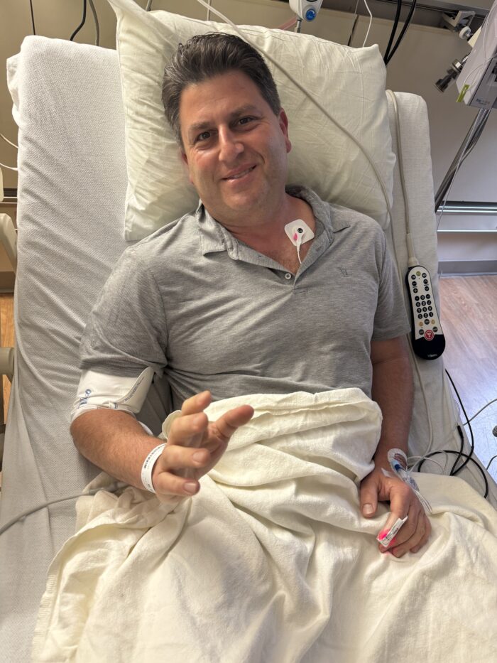 A man sits in a hospital bed with medical cords connected to his body and a white sheet pulled over his body. He is looking at the camera and smiling.