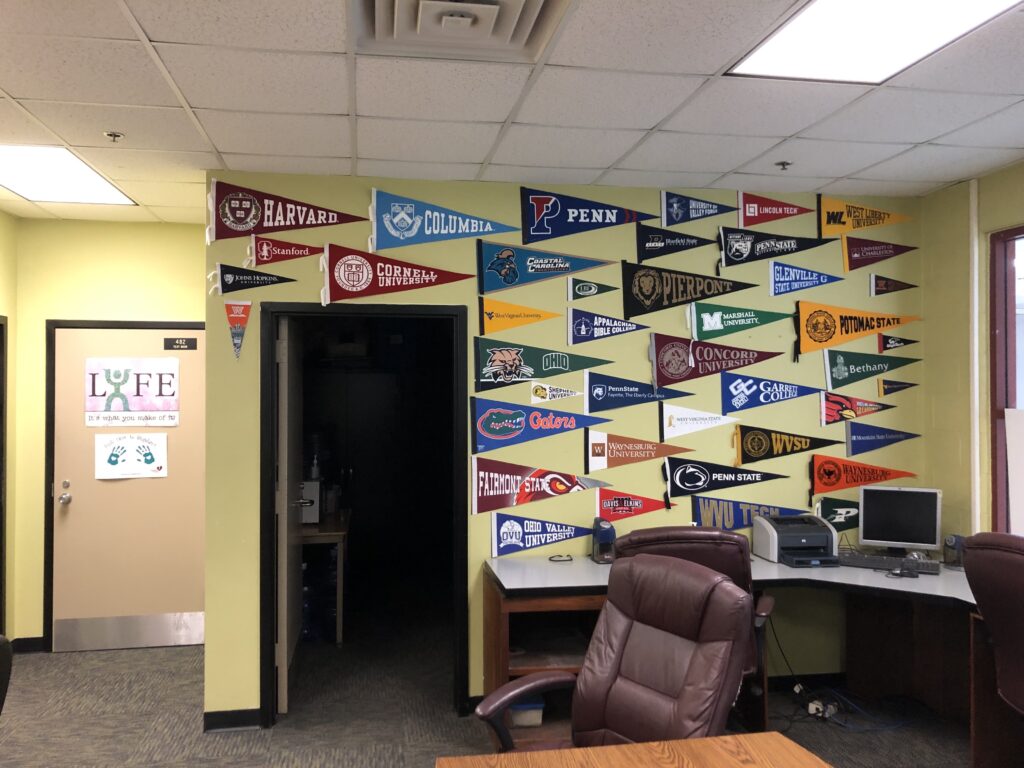 Pennants crowd a creme-yellow wall in what appears to be an office. There is the end of a wood table in the foreground with a plush office chair in front of it. The pennants on the wall include Harvard, Cornell, Penn, Columbia, Pierpont, and many other regional and national schools. There is a door to a dark office just left of the center of frame around which the pennants are arrayed.