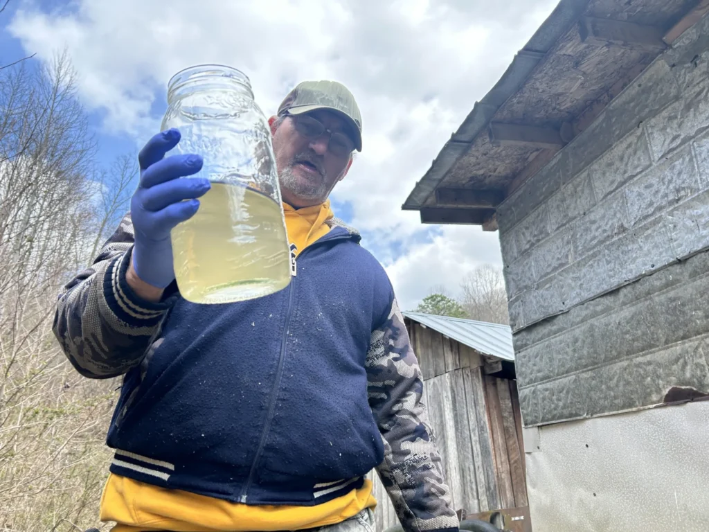 A man with blue, latex gloves on, holds up a jar of water. The water inside is murky and brown. The man wears glasses, a ball cap, and a blue jacket.