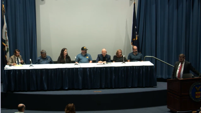 Seven people sit behind a long table covered in a blue tablecloth with microphones. A man wearing a suit and red tie stands to the right behind a wooden podium, conducting a panel discussion.