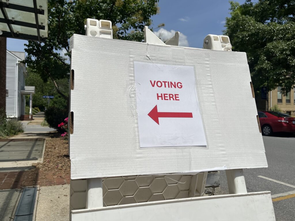 A white sign on a town street displays the words "Voting Here" and a large arrow pointing to the left. Sidewalks and parked cars are visible in the background.