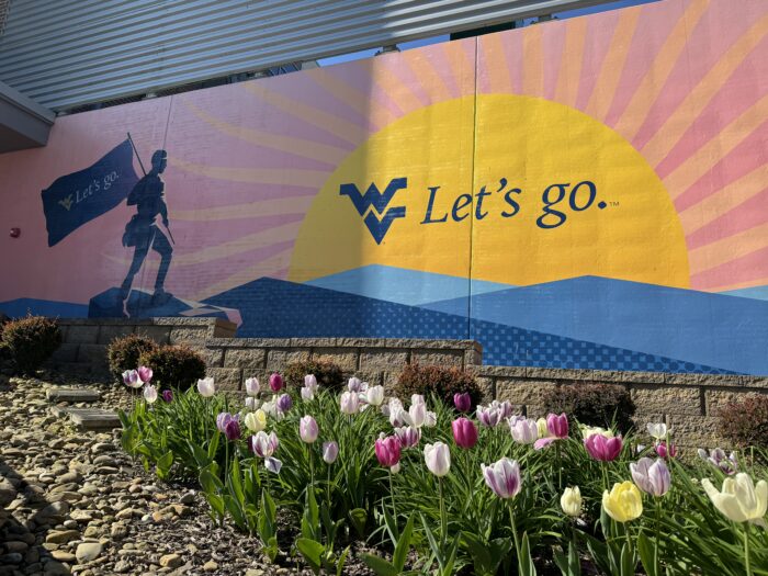 A bed of flowers is seen near the bottom of the photo. Above the flowers is a painted sign on a wall that features a sunrise and a silhouette of a mountaineer holding a flag. On the sun is the WVU logo and the words, "Let's go."