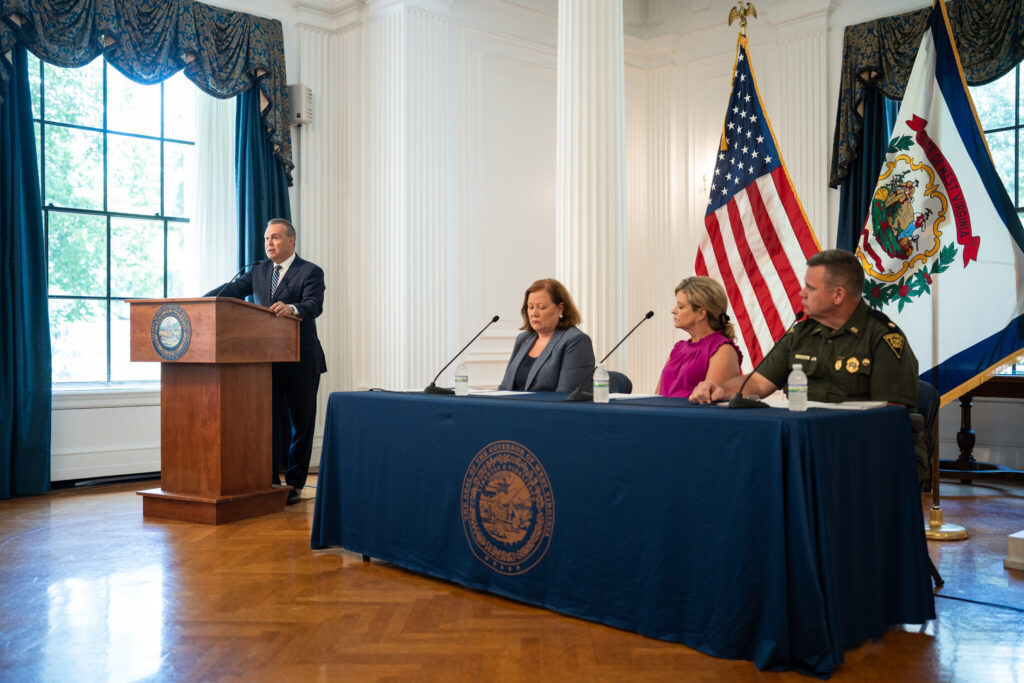 A man stands at a podium and three people sit at a desk with microphones Infront of them. They mood is serious.