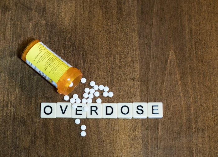 Tan tiles with black capital letters spelling "overdose" with an open prescription bottle and white tablets spilling out onto a wood table.