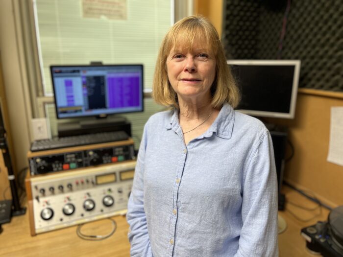 A woman stands in front of radio equipment, smiling for the camera.