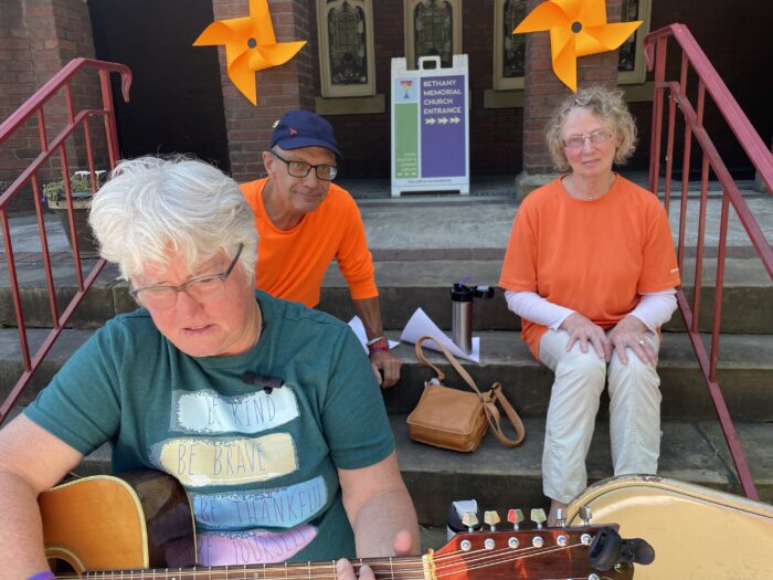 A woman holding a guitar sits on concrete steps. Behind her sit a man and a woman wearing orange shirts, smiling at the camera. In the background, orange pinwheels are placed on the exterior of a red brick building with a sign that says "Bethany Memorial Church Entrance." Further behind the sign is a row of small stained glass windows.