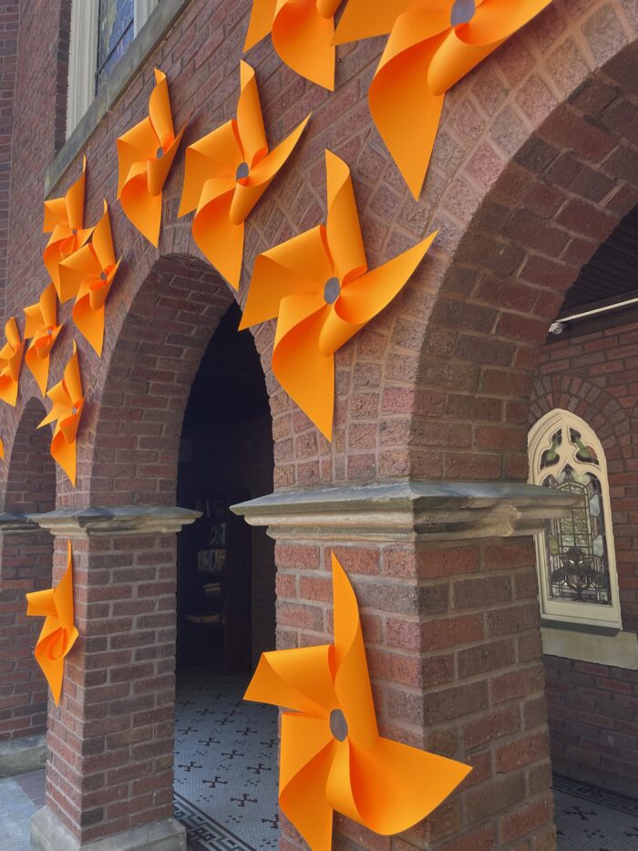 Orange pinwheels line the front of a brick church, covering three archways. Behind the archways are a sign and stained glass windows.