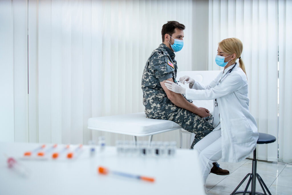 A male soldier with brown short hair in uniform sits atop an examination table as a female blonde physician injects the soldier with a vaccination. They are in a doctor's office with all white furnishings and wearing blue masks.
