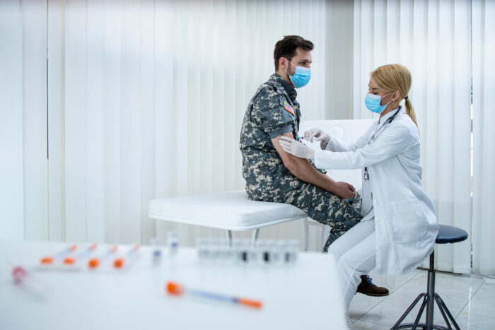 A male soldier with brown short hair in uniform sits atop an examination table as a female blonde physician injects the soldier with a vaccination. They are in a doctor's office with all white furnishings and wearing blue masks.