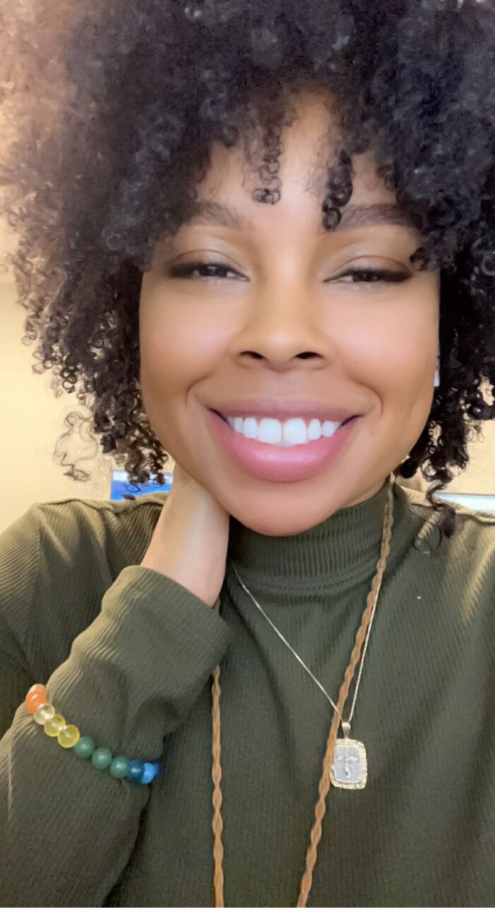 A selfie of a smiling woman with dark hair and dangling earrings. She wears an olive turtleneck shirt.