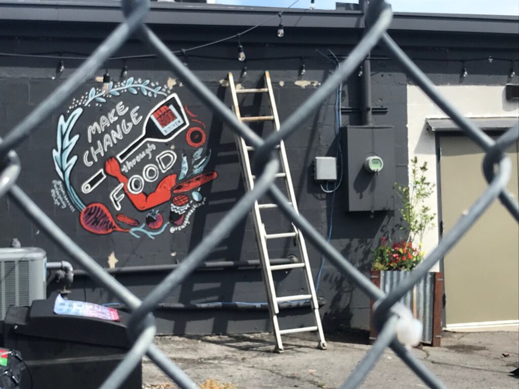 The outside of a restaurant. Close to the camera is a chain link fence. Beyond the fence, painted on the side of the restaurant, are the words, "Make Change through Food."