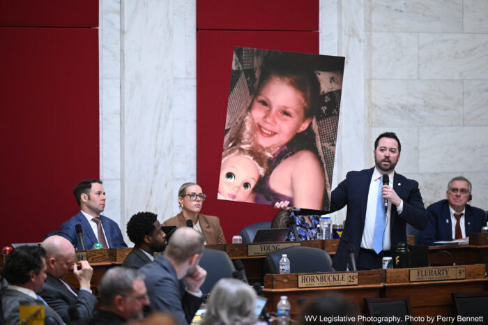 A man in a suit, amongst other legislatures holds a sign of a little girl in one hand, and a microphone in another.