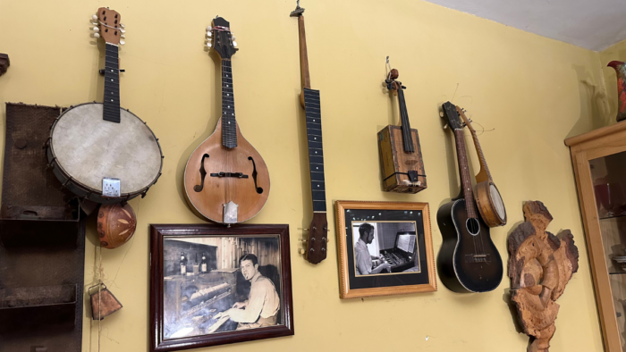 A variety of instruments are seen hung on the wall. Instruments, such as a mandolin, guitar, fiddle, etc. Underneath the images are black and white photographs.