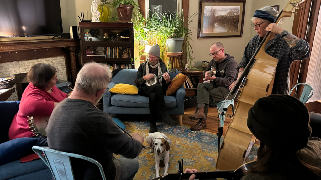 Six people sit in a circle together in a room in a home. They each have a different instrument. In the center of the circle is a dog resting.