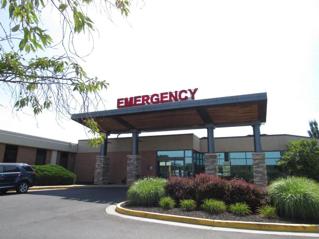 A short building with an outstretched roof bears a sign with the word "Emergency" on it in bright red letters. In front of the building is an asphalt driving loop with a small garden in the middle. A car is partially visible off to the side.