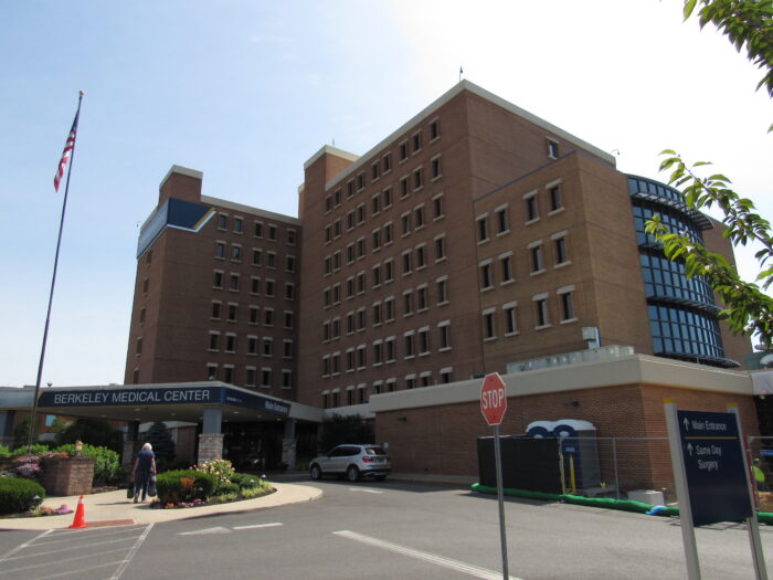 A tall brick building with may windows and an American flag out front displays a sign that says "Berkeley Medical Center." A car is pulled in front of it, and a person is walking on the sidewalk toward the entryway.