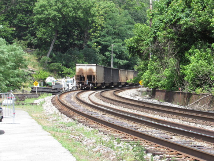 A train travels along railroad tracks, surrounded by trees with green leaves. The train is visible from the back, with the caboose traveling away. A sidewalk is also visible on its side.