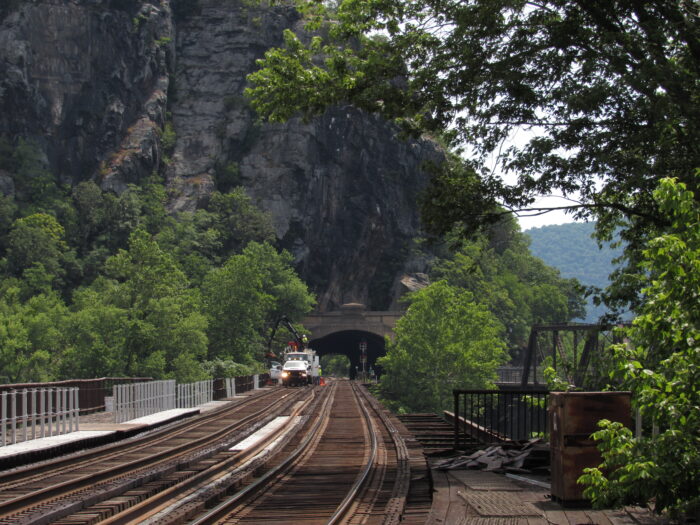 Railroad tracks lead into a tunnel carved into a mountainside. Trees with green leaves surround the tracks on either side. A car is parked on the railroad tracks in the distance, with men in neon yellow garments beside it.