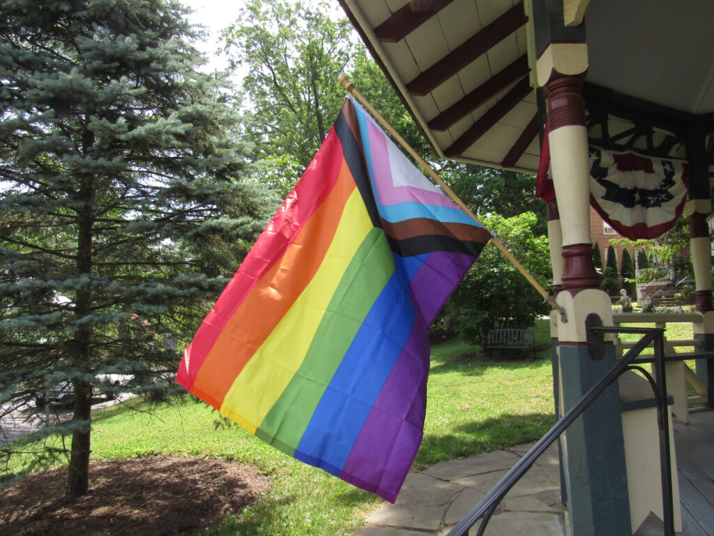 A rainbow flag with a triangle on the side near the flagpole that contains white, pink, blue, brown and black stripes. The flag waves in the wind, mounted on the post of a gazebo.