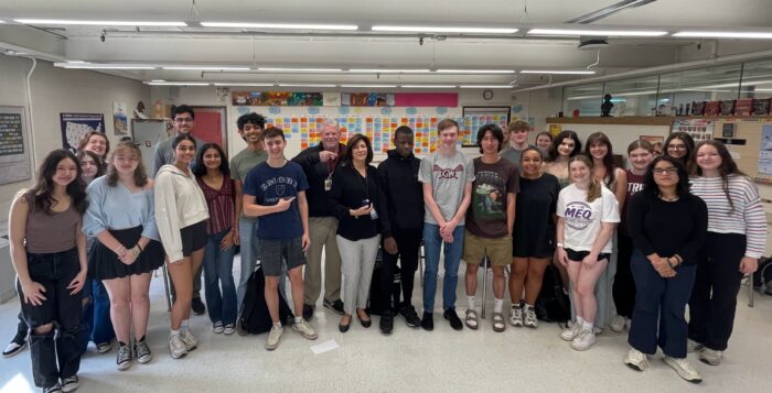 Twenty-four people are seen in a group photo in a classroom. Two adults and 22 teenagers. They are all dressed casually and comfortably. In the center of the group are the two adults - a middle age woman with brown hair wearing a black blouse and light colored slacks. Behind her is the second adult, a man, with white hair. He smiles and points to the woman next to him.