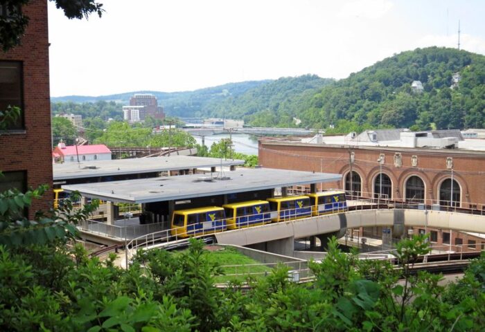 A landscape frame shows a rounded, forested hill dominating the top right of frame. Below the hill can be seen a red brick building in the midground, with what appears to be a rail station in the foreground, with foliage in the extreme foreground. The station has four cars parked at it, all four in a blue and yellow color scheme, with the sides facing camera in blue with a WV logo in yellow. The left of frame is dominated by a brick wall with windows. In the background right of frame can be seen a bridge spanning a body of water, the Monongahela River, and further back a small skyline that leads into a hill that slopes right into the rounded hill mentioned above.