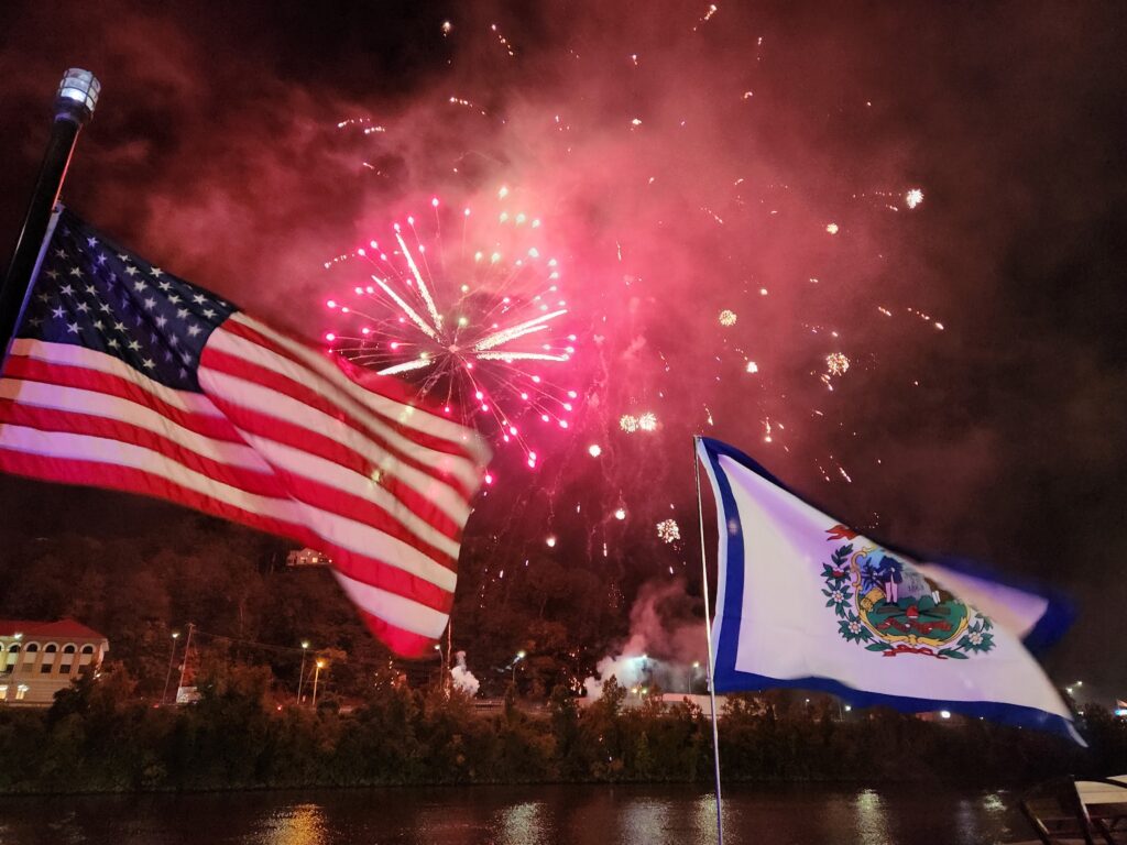 firework shells explode over the river with flags in the foreground