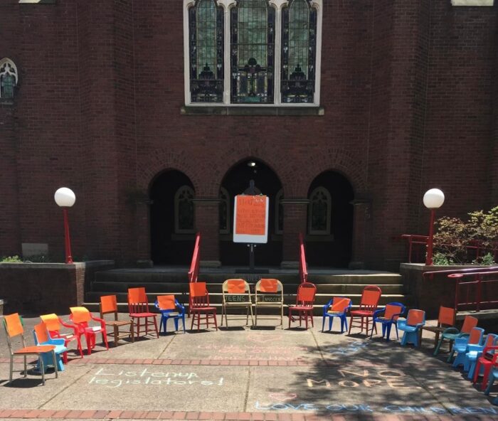 Twenty-one chairs with orange signs sit before a church entryway on a sidewalk.