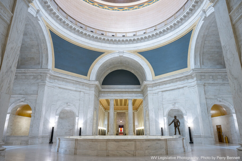 A wide shot of the center of the Capitol Rotunda from the second floor. There are white, marble walls, and the room is well lit. There is a statue of Robert C. Byrd in the distance.