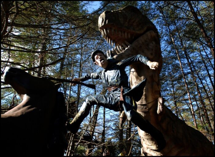 A photograph of a Civil War soldier statue being carried by a t-rex dinosaur. The statue of the soldier appears as if it were lifted off of a horse.