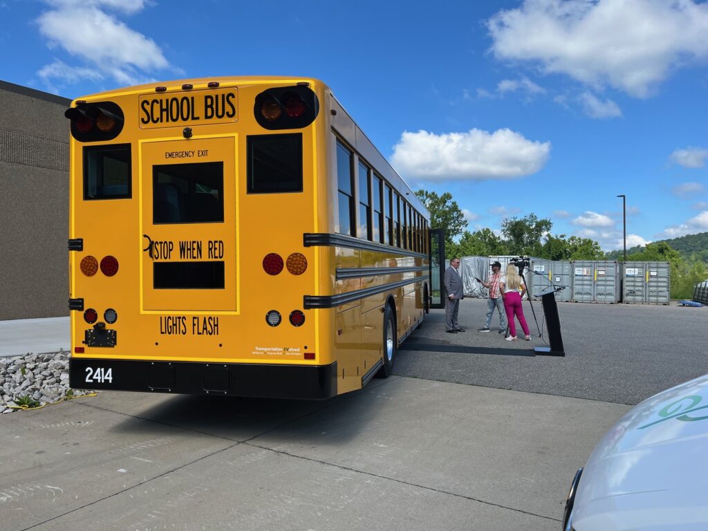 A yellow school bus, powered by electricity sits in the lot outside the factory where it was built as a company official talks to a TV news crew with a blue sky overhead on a summer day.