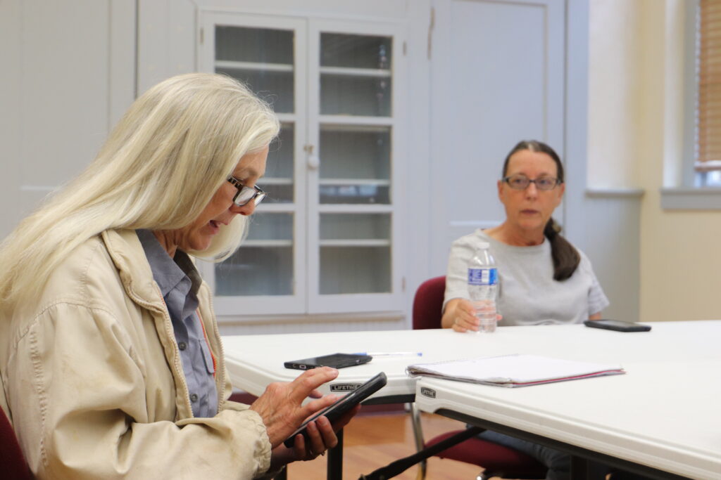 Two women sit at a table in a classroom in a converted schoolhouse. One is looking at her phone, the other is looking straight ahead. One has long light hair, the other has dark hair in a ponytail.